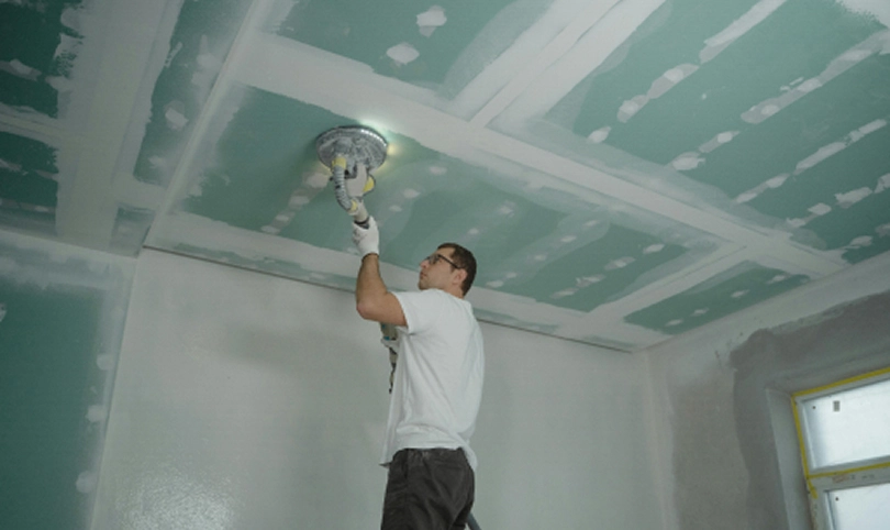 Person sanding a drywall ceiling with an electric sander in a room under renovation.