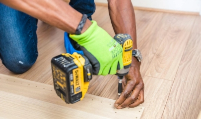 Person using electric screwdriver on wooden floor, wearing green gloves; close-up on hands and tool.
