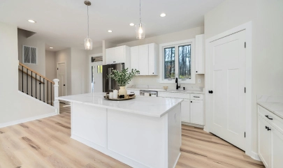 Modern white kitchen with island, wooden floors, pendant lights, and large window. Sleek design with ample storage.