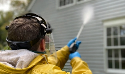Person pressure washing house exterior siding, wearing protective gear and gloves for home maintenance cleaning.