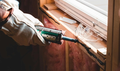 Person sealing a window frame with caulk, wearing gloves for insulation installation in a wooden structure.