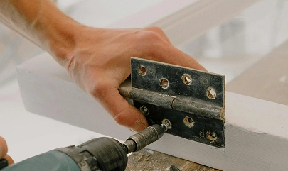 Person using a power drill to attach a hinge on a white wooden frame.