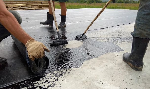 Workers applying waterproofing membrane to a concrete surface using rollers and protective gear outdoors.