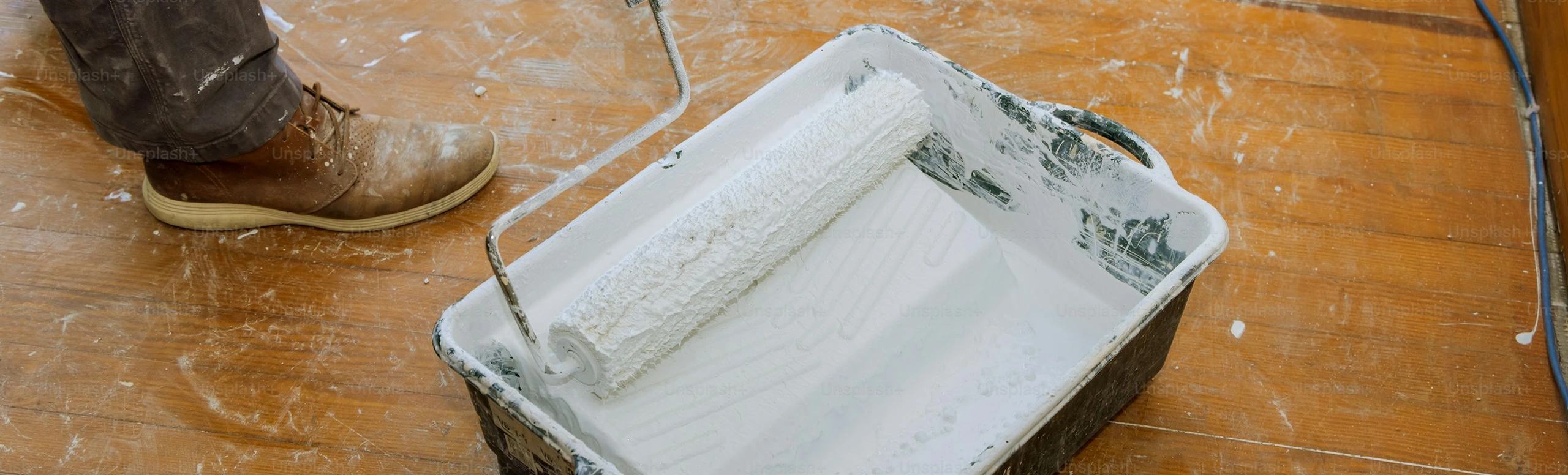 Paint roller in tray with white paint on wooden floor, next to person wearing brown boots.