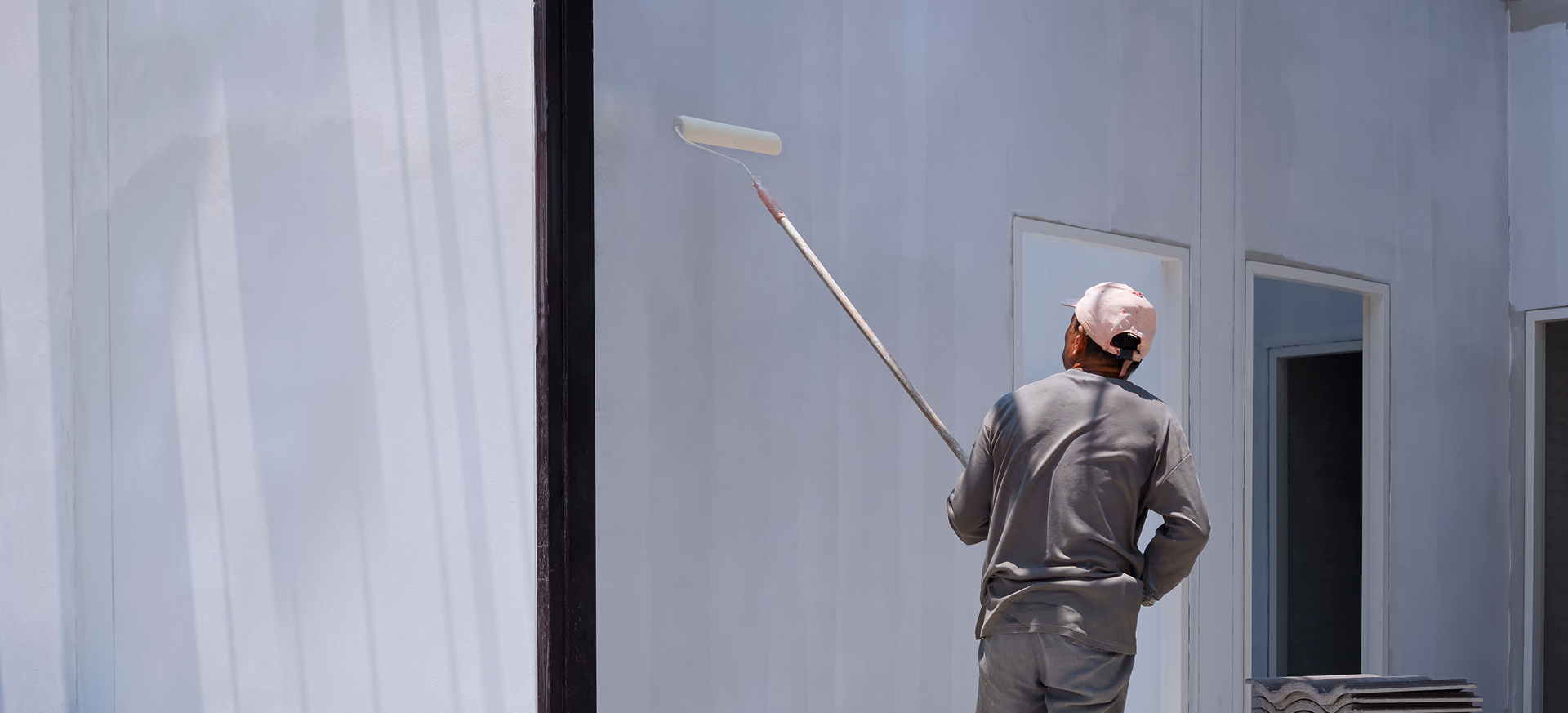 Painter using a long-handled roller on a light grey exterior wall, seen through a dark steel door frame on a sunny site